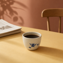 Keramik kopper med blå bier og kaffe på gul bordplade med avis og stol