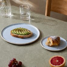 Keramik tallerkener lyseblå med avokadomad og croissant og frugt på et bord med stol 