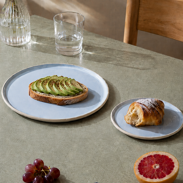 Keramik tallerkener lyseblå med avokadomad og croissant og frugt på et bord med stol 