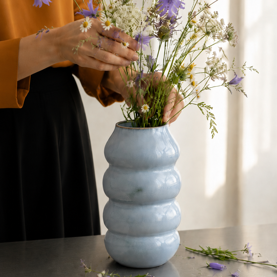 Vase i stentøj med bløde former drejet i hånden, lyseblå. Kvinde sætter blomster i vasen. 