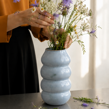 Vase i stentøj med bløde former drejet i hånden, lyseblå. Kvinde sætter blomster i vasen. 