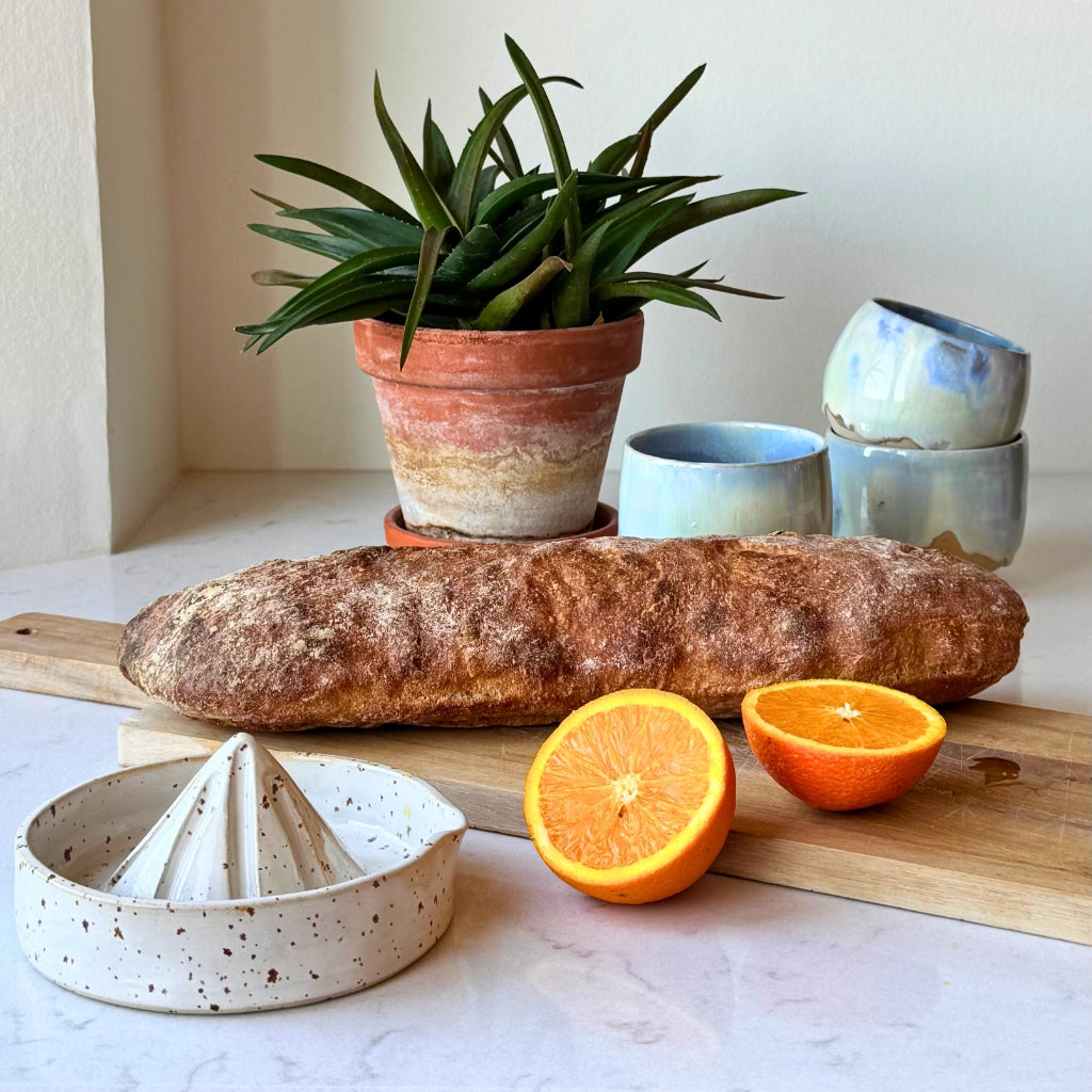 Citruspresser i keramik på lys bordplade med opstilling med appelsiner, brød og keramikkopper.