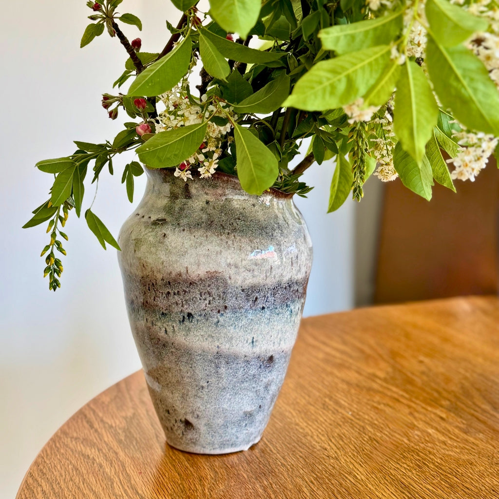 Keramik vase til blomster med grønne grene og hvide blomster på træbord.