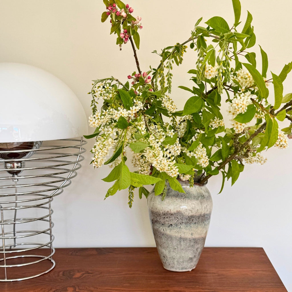 Keramik vase til blomster med grønne grene og hvide blomster på træbord med designerlampe