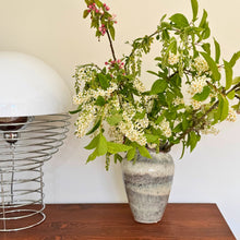 Keramik vase til blomster med grønne grene og hvide blomster på træbord med designerlampe