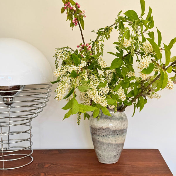 Keramik vase til blomster med grønne grene og hvide blomster på træbord med designerlampe